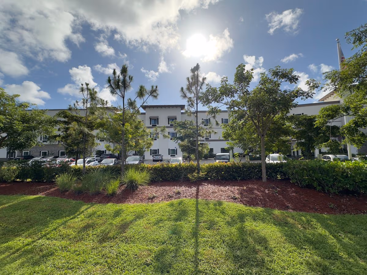 Front exterior of a three-story building with lawn, trees, and parked cars under a sunny sky.