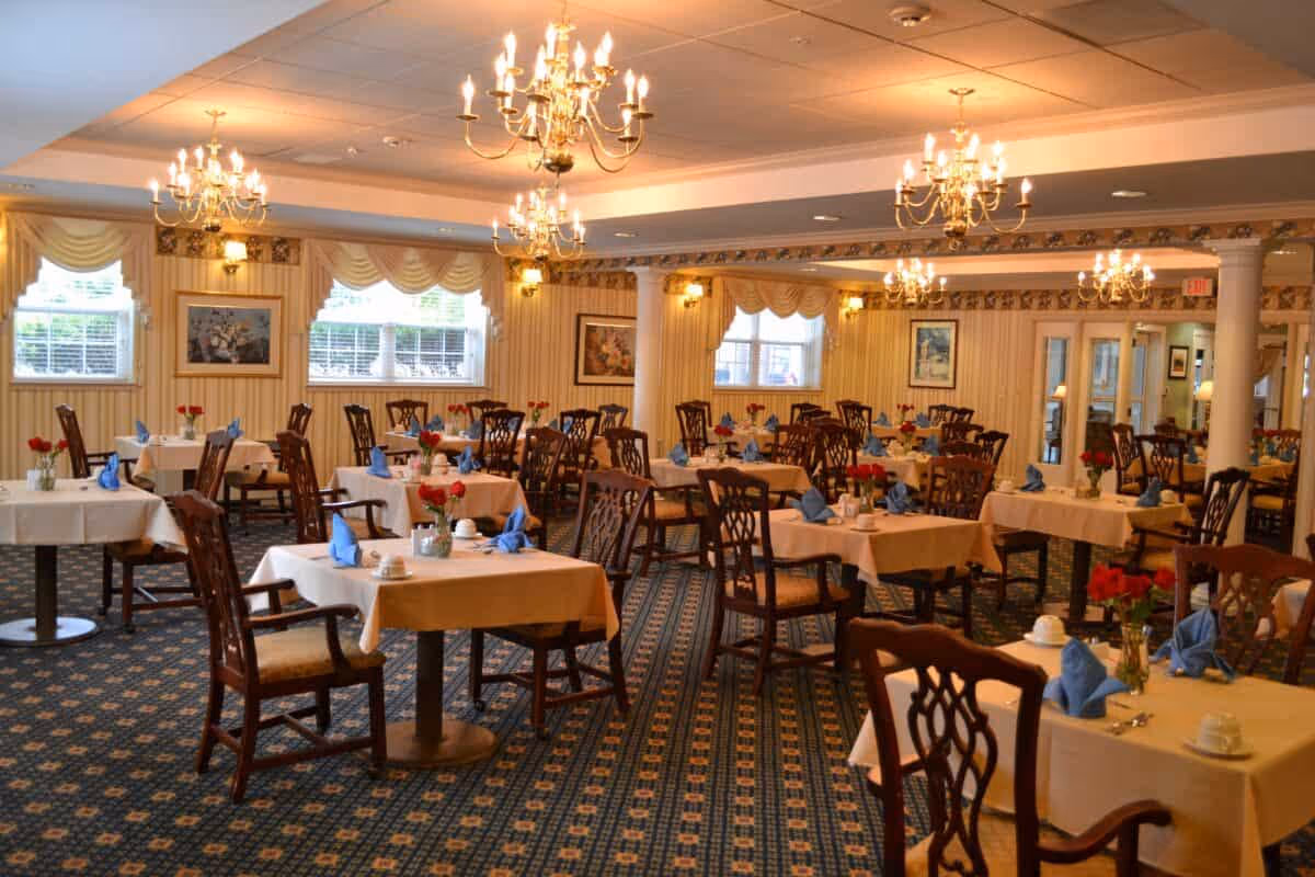 A well-lit dining room with multiple tables covered in white tablecloths, each set with blue folded napkins, cups, and small vases of red flowers. The room features patterned carpet, chandeliers hanging from the ceiling, and windows with draped curtains. The walls are decorated with framed artwork.