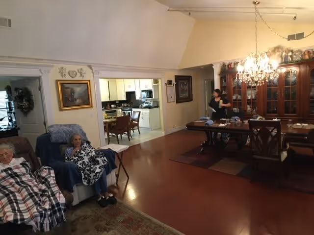 A cozy living and dining area in a senior living facility. Two elderly women are seated on armchairs with blankets, and a caregiver is standing near a large wooden dining table with chairs. The room has hardwood floors, a chandelier above the dining table, and a china cabinet filled with decorative items. In the background, a kitchen area is visible.