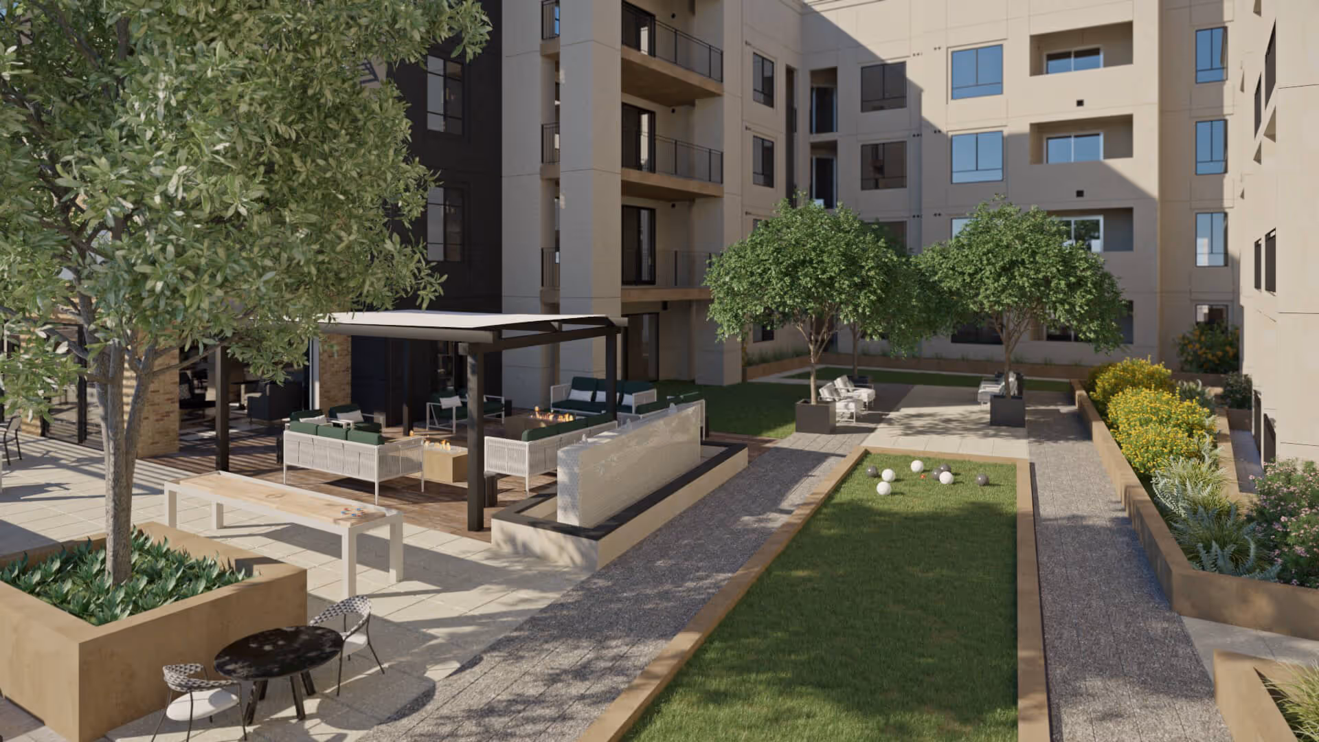 Outdoor courtyard area of a senior living facility with green trees, a bocce ball court, seating areas including a shaded lounge with sofas and fire pits, and a small table with chairs. The courtyard is surrounded by a multi-story building with balconies and windows.