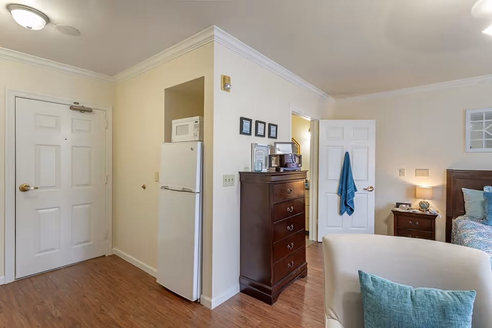 Interior view of a senior living facility room showing a white entrance door, a small kitchen area with a white refrigerator and microwave, a dark wooden chest of drawers with framed pictures and a radio on top, an open door with a blue towel hanging on the handle, a bedside table with a lamp and a bed with blue and white bedding, and a beige armchair with a blue cushion in the foreground.