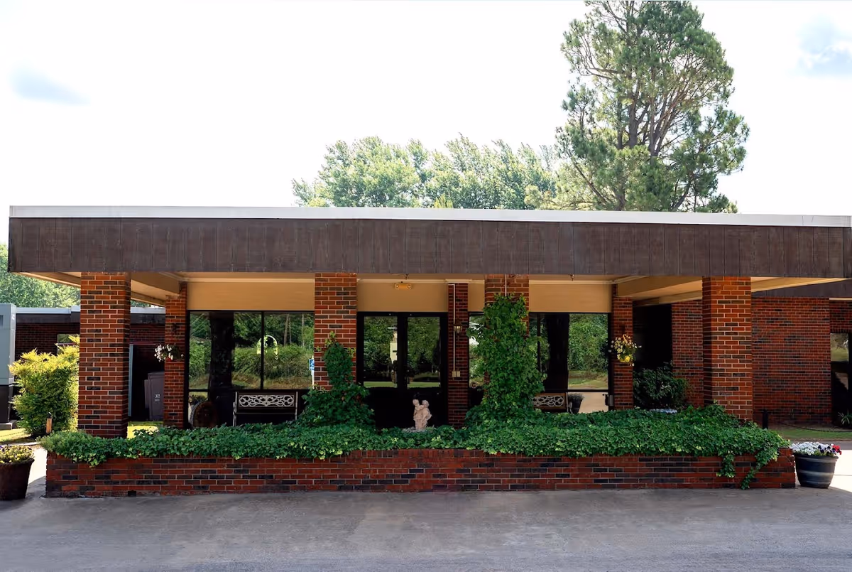 Front exterior view of Stilwell Nursing & Rehab building with brick pillars, large windows, and greenery including bushes and potted plants.