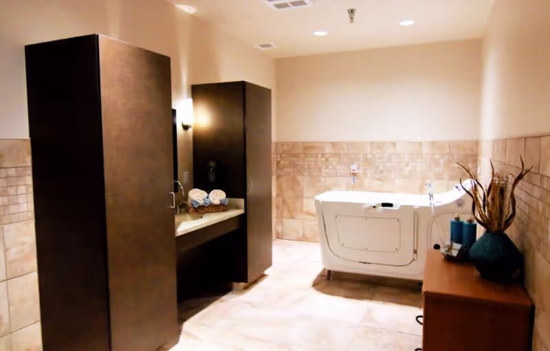 A spacious bathroom with beige tiled walls and floor, featuring a walk-in bathtub on the right side, a wooden cabinet with decorative items, and a sink area with rolled towels and dark wood cabinets on the left.