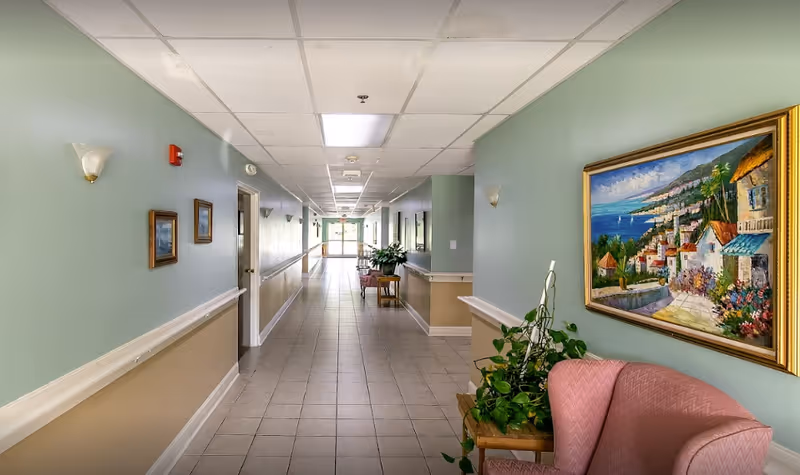 A long, clean hallway in a senior living facility with light green and beige walls, tiled floor, ceiling lights, framed paintings on the walls, potted plants, and pink upholstered chairs along the sides.