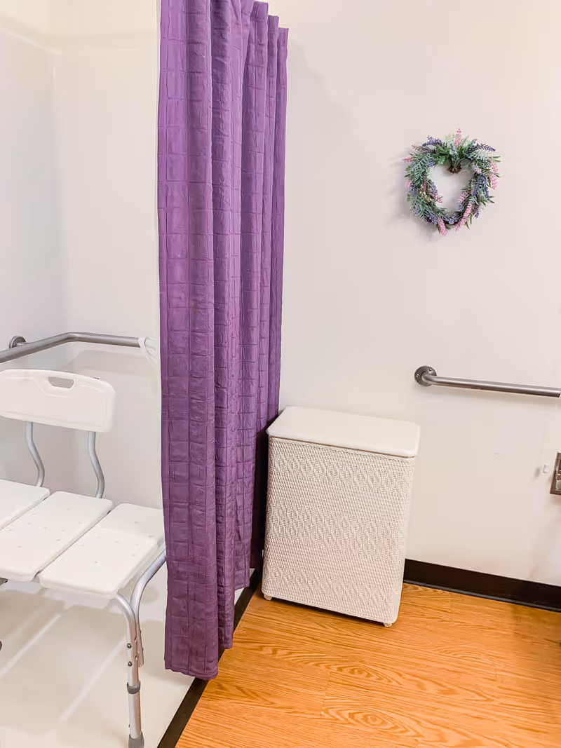 A corner of a bathroom with a white shower chair, a purple shower curtain, a white woven laundry hamper, a metal grab bar on the wall, and a decorative heart-shaped wreath hanging on the wall. The floor is wood-patterned.