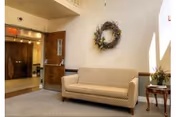 A small seating area with a beige patterned couch against a light-colored wall. Above the couch is a decorative wreath. To the right of the couch is a small wooden side table with a floral arrangement. An open door leads to a hallway with carpeted floors and light walls.