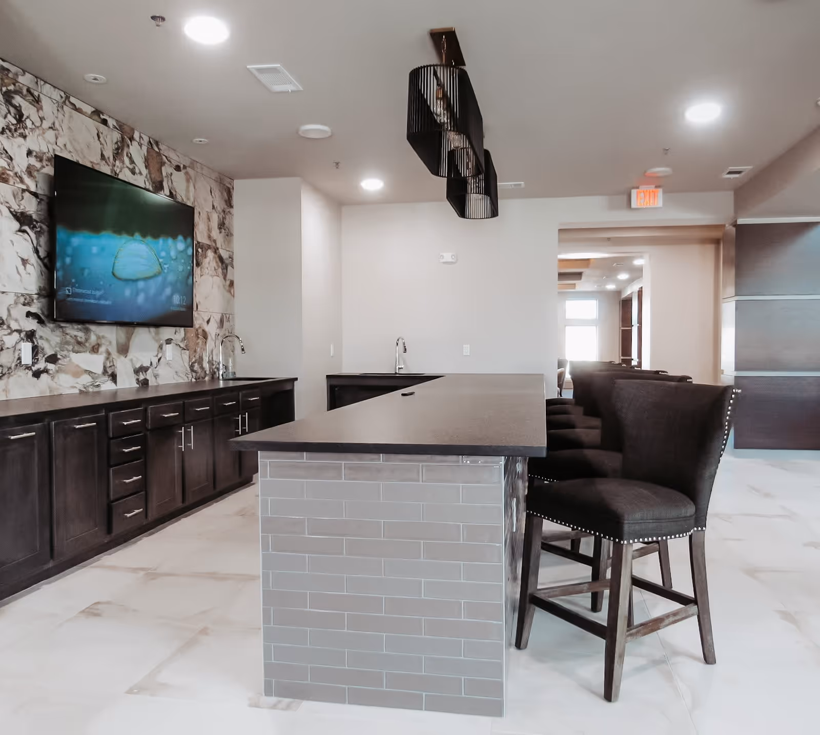 Modern interior space featuring a long island counter with dark countertop and gray tiled base, surrounded by high-back dark upholstered chairs. The back wall has dark wooden cabinets with silver handles, two sinks, and a mounted flat-screen TV displaying an image of a water droplet. The floor is light-colored tile, and the ceiling has recessed lighting and a unique black pendant light fixture.