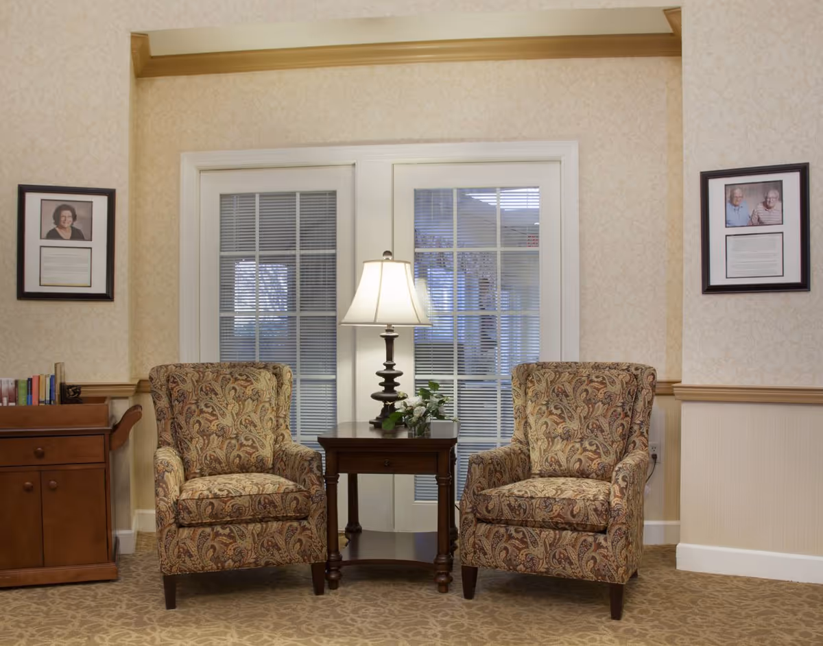 A cozy sitting area with two patterned armchairs placed on either side of a wooden side table with a lamp and a small plant. Behind the chairs are white French doors with glass panes and blinds. The walls are decorated with light patterned wallpaper and two framed pictures with text are hung on either side of the doors. A wooden cabinet with books is visible on the left side.