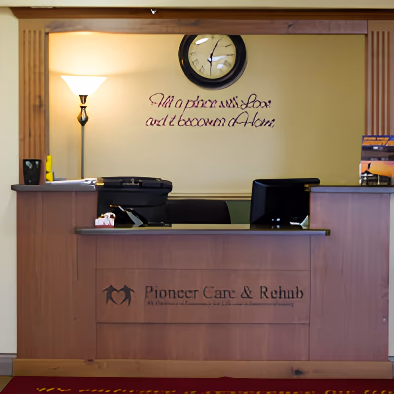 Reception desk in a senior care facility lobby with a wall clock, floor lamp, two monitors, and 'Pioneer Care & Rehab' signage on the front desk.