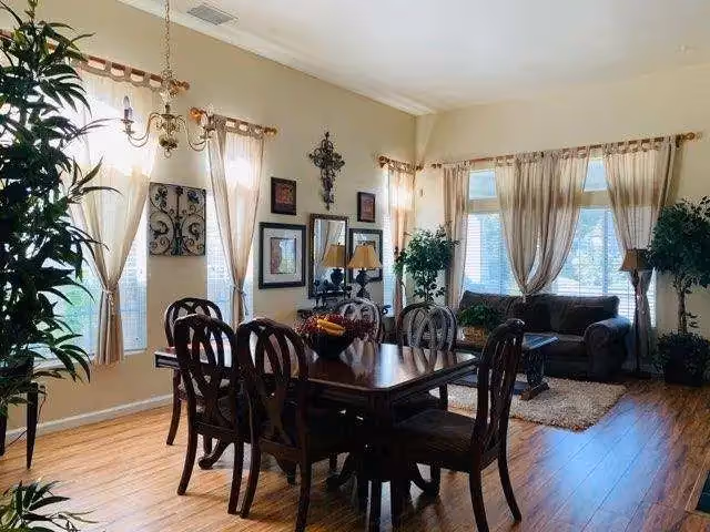 Sunlit open dining and living area with a wooden dining table and chairs, sofa, plants, wall art, and large curtained windows.