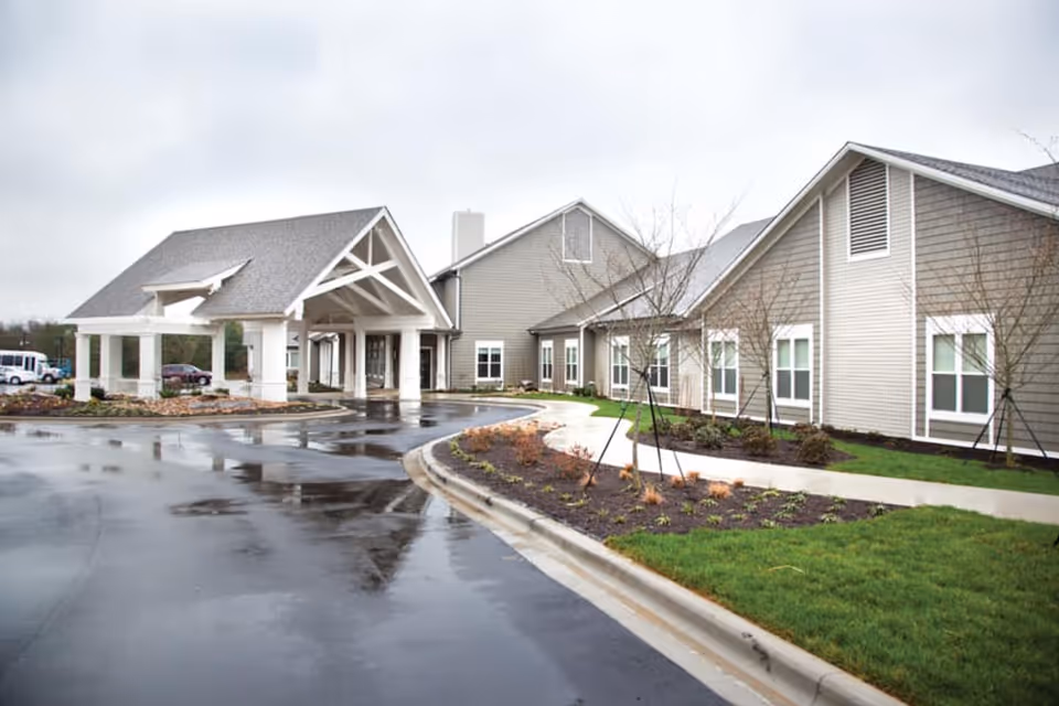 Exterior view of a senior living facility building with a covered entrance, wet pavement, landscaped garden beds, and young trees on a cloudy day.