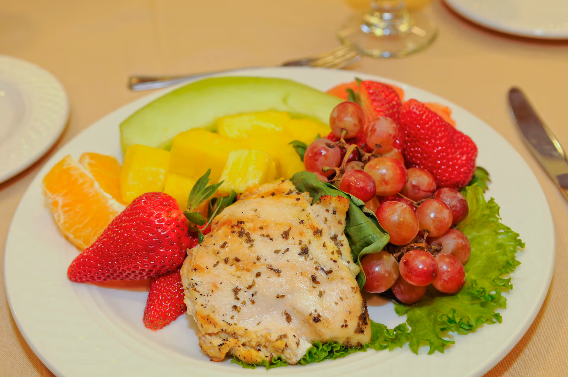 A plate with grilled chicken served on lettuce alongside strawberries, grapes, pineapple, melon, and orange segments.