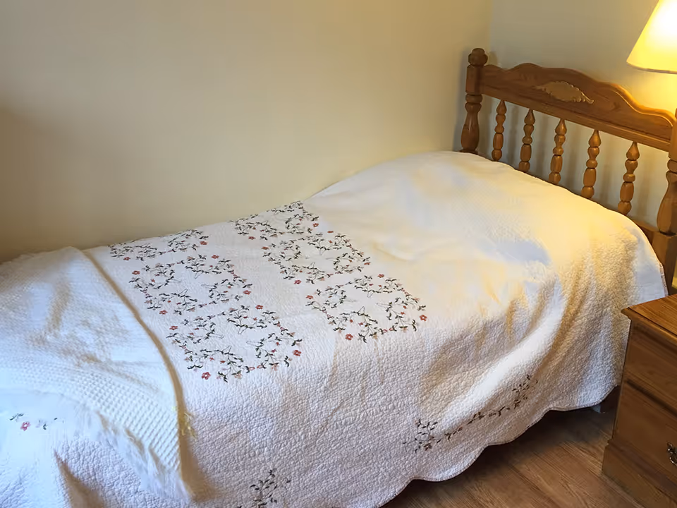 A single bed with a wooden headboard covered by a white floral quilt next to a wooden nightstand and lamp.