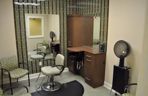 Interior view of a small salon or grooming area with a styling chair in front of a mirror, a cabinet with drawers and a sink, two additional chairs with patterned upholstery, and a hair dryer on a stand in the corner.