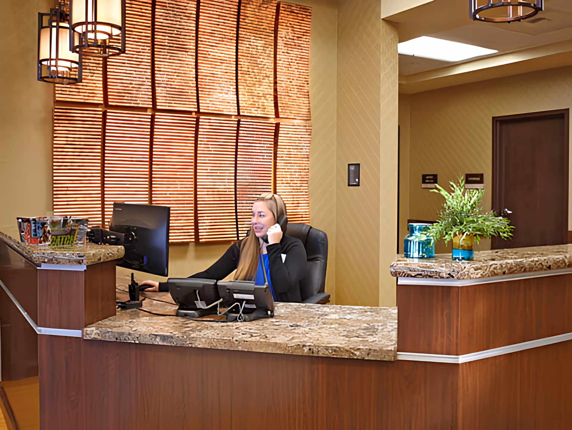 A receptionist sits at a marble-topped front desk in a warmly lit lobby with wooden blinds and decorative plants.