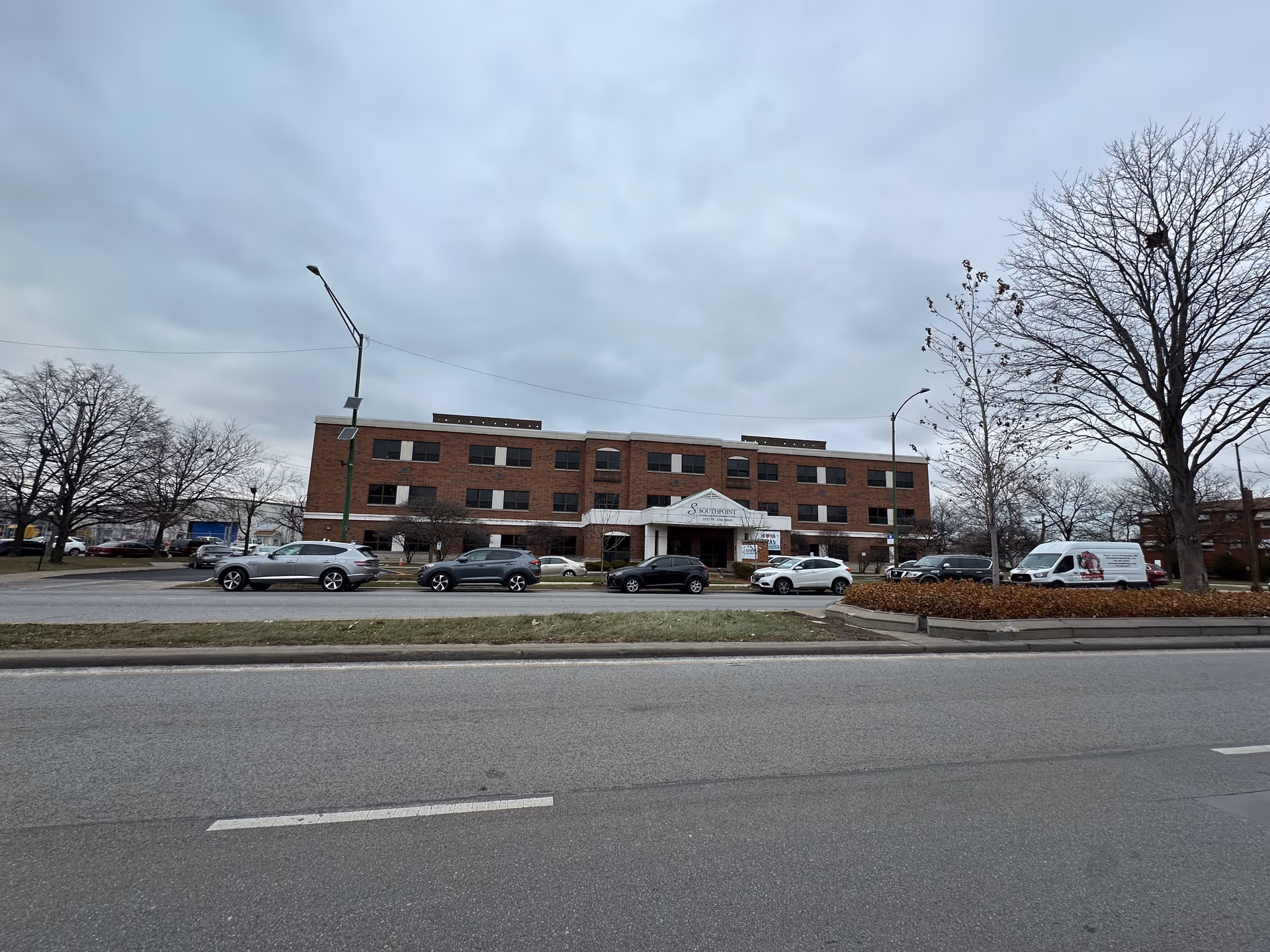 Exterior view of a three-story brick building with multiple windows, identified as Southpoint Nursing and Rehabilitation Center. Several cars are parked in front of the building along the street. Leafless trees and a cloudy sky are visible.