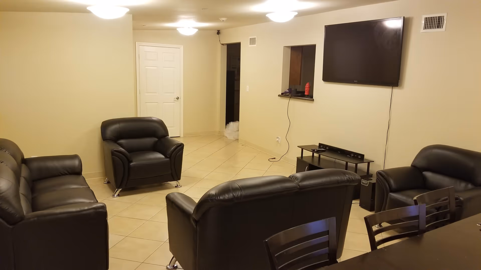 A living room with beige walls and tiled floor featuring black leather sofas and armchairs arranged around a wall-mounted flat screen TV. There is a small black TV stand below the TV with electronic devices on it. A dark wooden dining table with chairs is partially visible in the foreground.