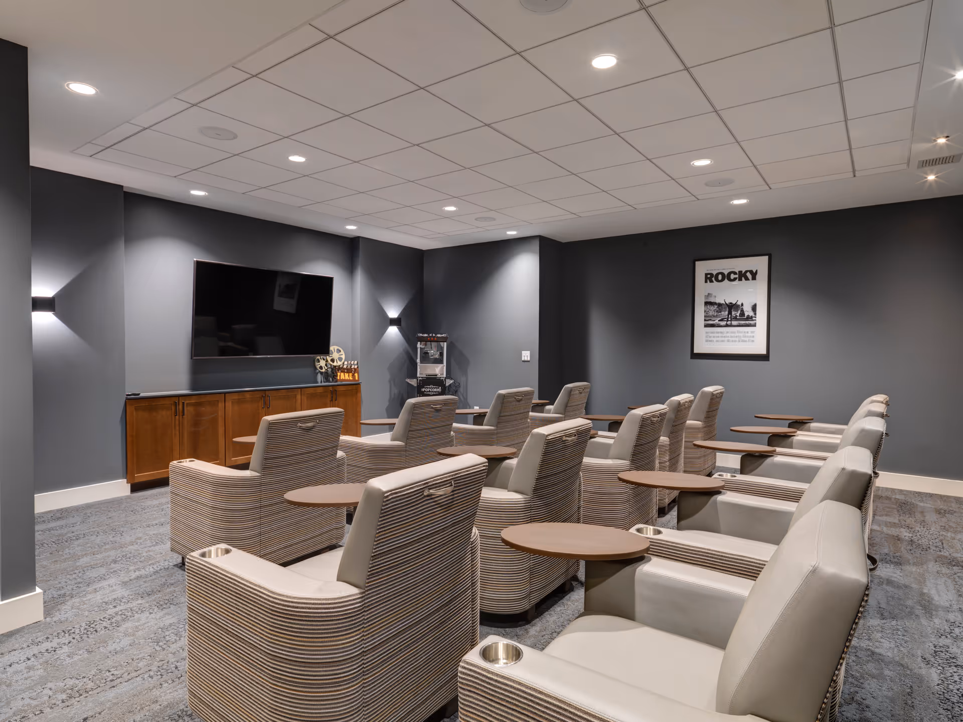 A small theater room with two rows of comfortable beige armchairs, each equipped with a wooden side table and cup holder. The room has gray walls, a large flat-screen TV mounted on the front wall above a wooden cabinet, and a popcorn machine in the corner. A framed poster of the movie Rocky is displayed on the right wall. The ceiling has recessed lighting.