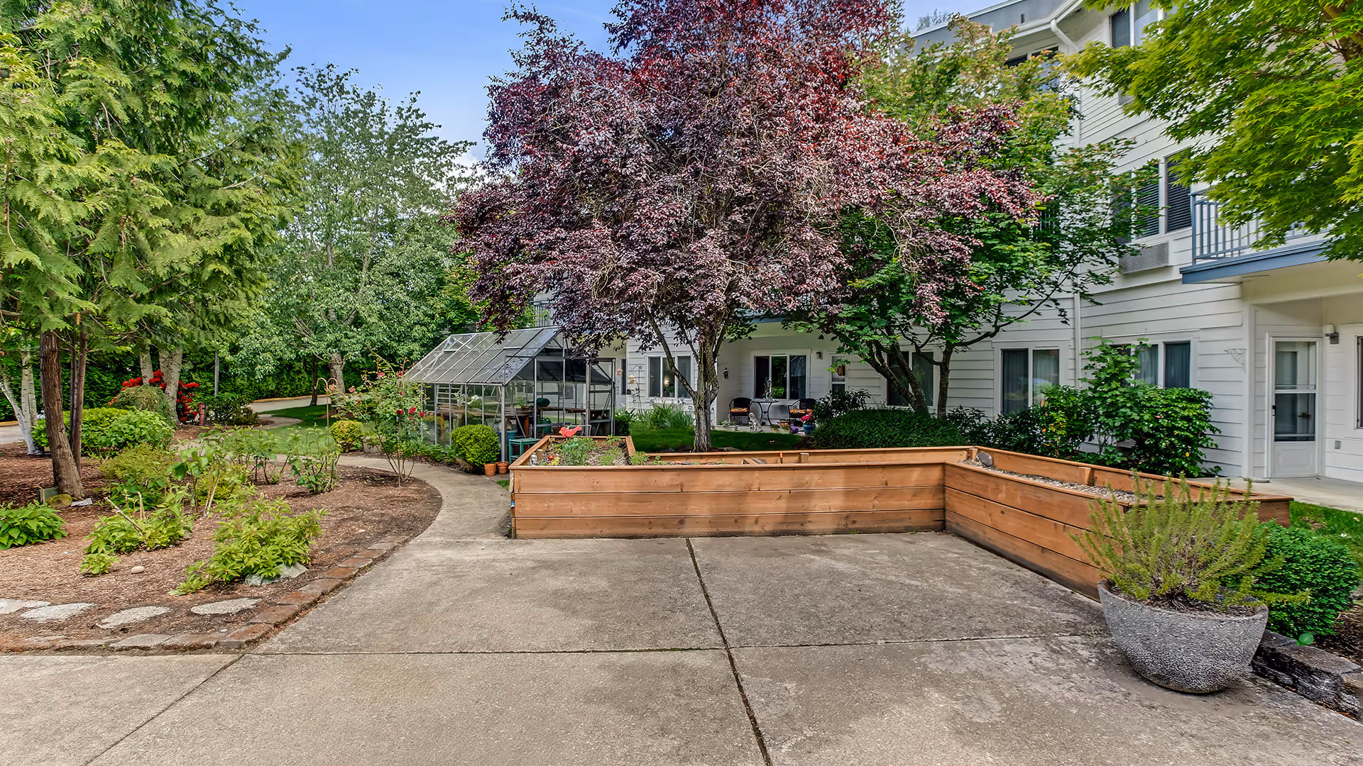 Outdoor garden area at Holiday South Hill featuring a concrete pathway, raised wooden garden beds, a small greenhouse, various trees and shrubs, and a multi-story building with balconies in the background.