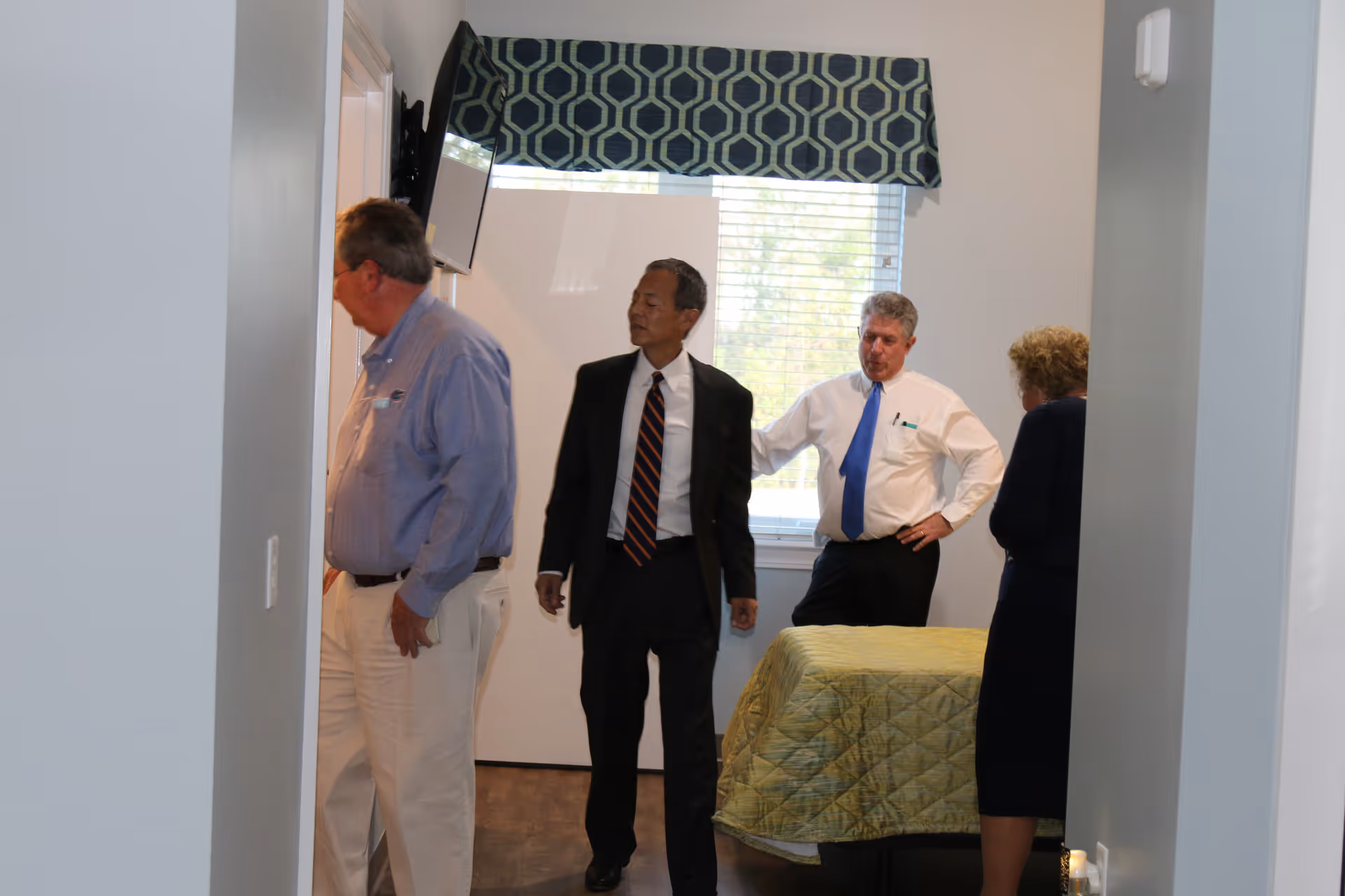 Four adults standing in a bedroom with a bed covered by a green quilt. The room has a window with blinds and a patterned valance. A wall-mounted TV is visible on the left side.