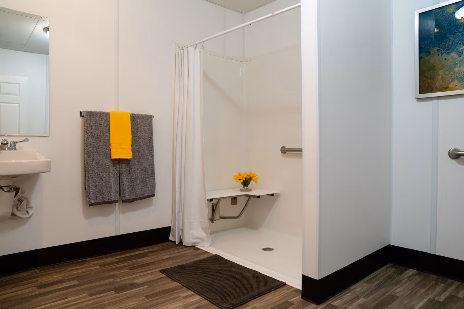 A clean and accessible bathroom with a walk-in shower featuring a fold-down seat holding a small vase of yellow flowers. There is a white shower curtain, a gray bath mat on the wood-patterned floor, a sink with a mirror above it, and two gray towels with a yellow towel hanging on a towel rack. A framed abstract artwork is visible on the wall to the right.