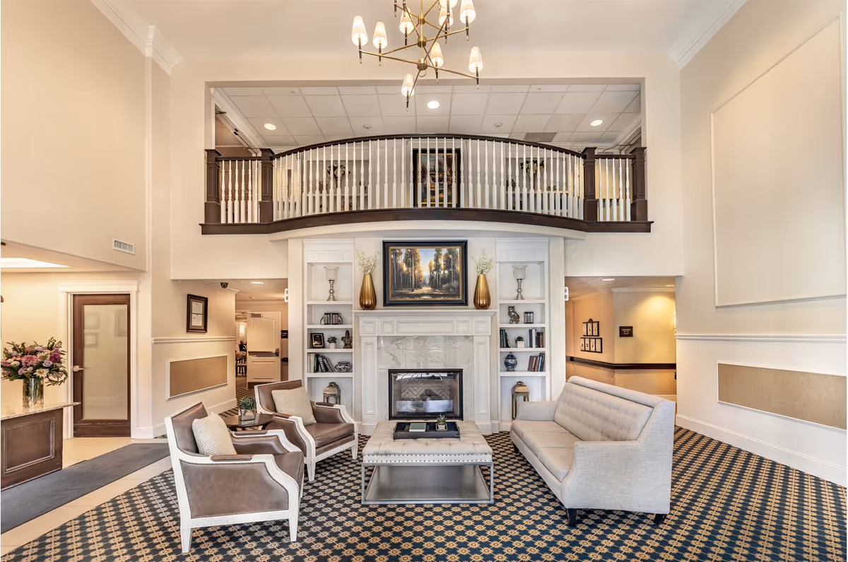 Elegant senior living lobby with chairs and a sofa arranged around a central fireplace and a balcony above.