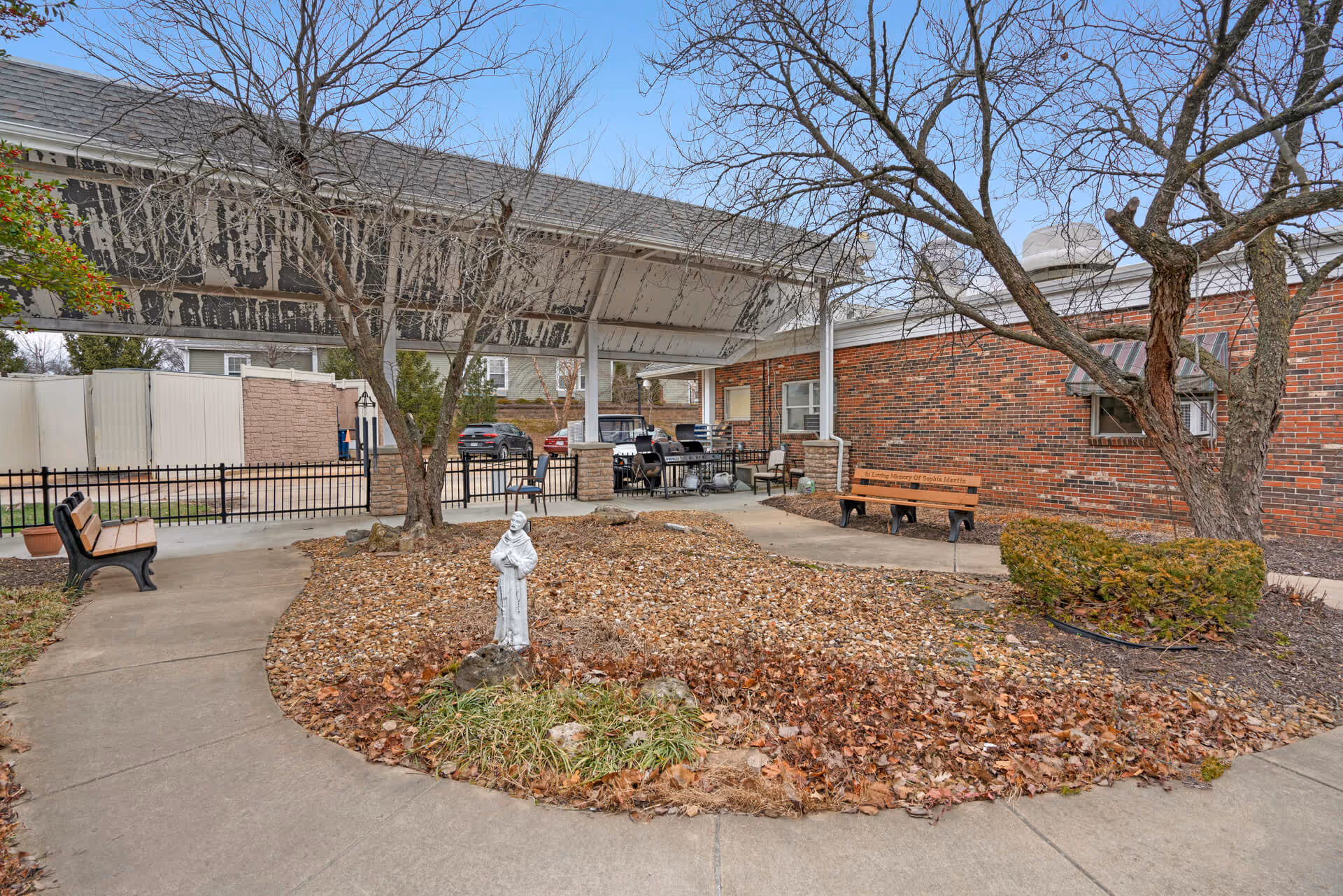 Outdoor courtyard area at StoneBridge Senior Living - Maryland Heights featuring a covered patio with seating and a grill, leaf-covered garden bed with a small statue, bare trees, and benches along the concrete pathways. The building exterior is brick with windows and awnings.