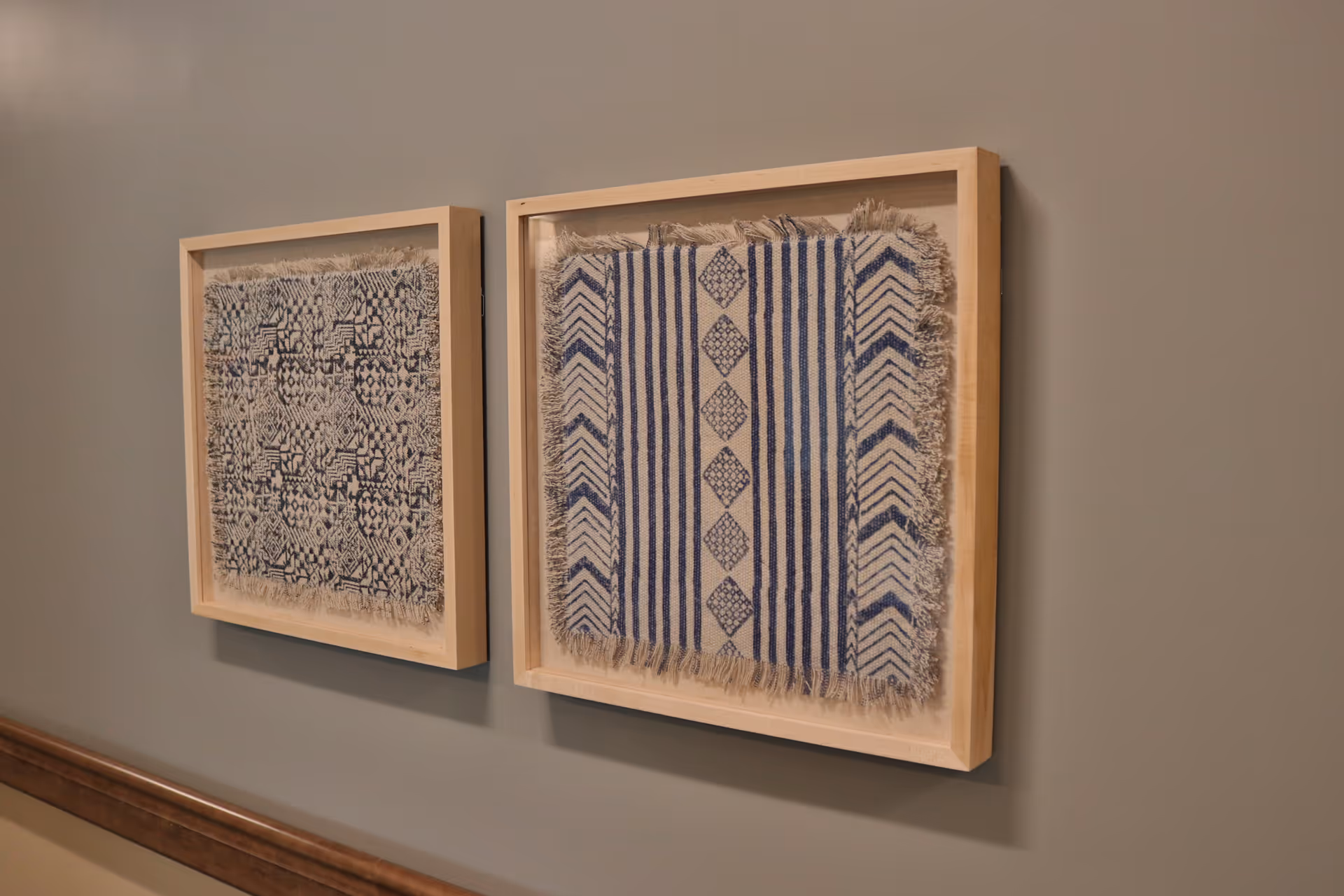 Two framed textile art pieces with blue and white geometric patterns hanging on a gray wall above a wooden chair rail.