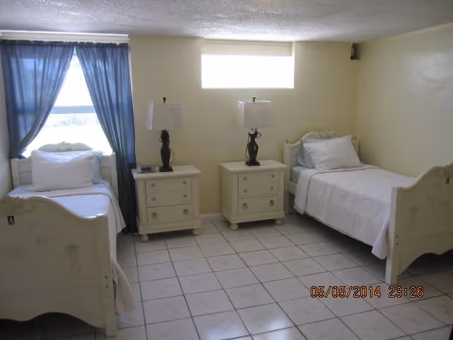 Small bedroom with two twin beds, two nightstands with lamps, a tiled floor, and a curtained window.