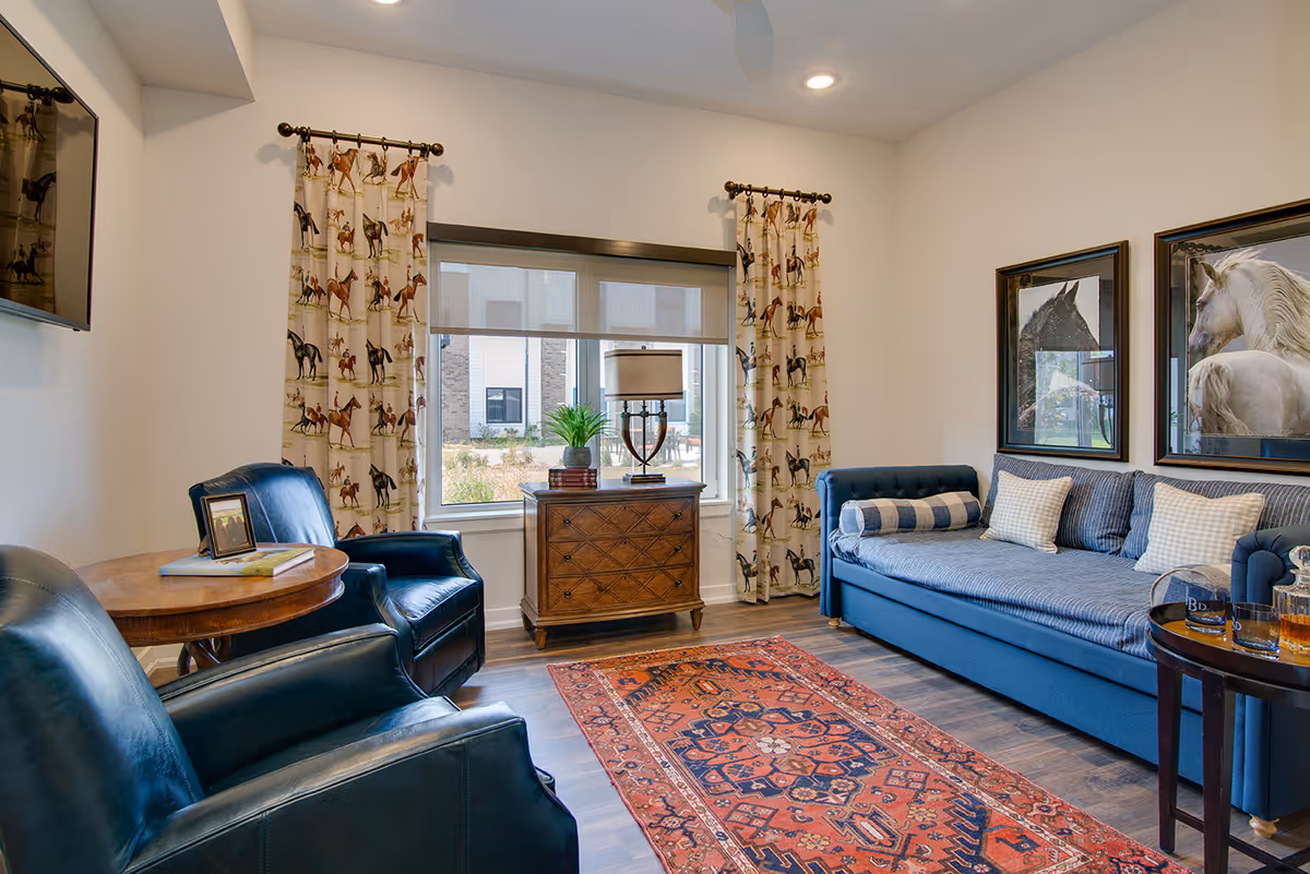 A cozy living room with a blue sofa adorned with multiple pillows, two dark blue leather armchairs, a wooden side table with books and a framed photo, a wooden chest with a lamp and a plant in front of a window with horse-patterned curtains, and two framed horse artworks on the wall. A red patterned rug covers part of the wooden floor.