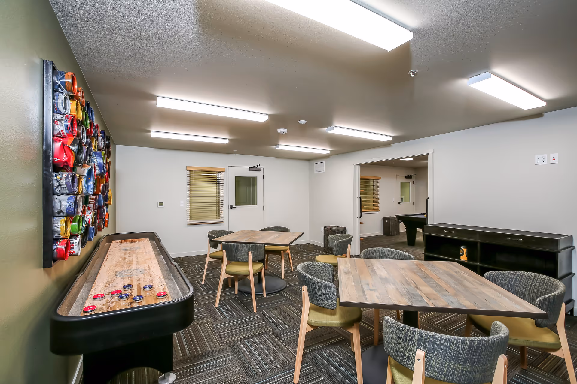 A recreational room with two wooden tables surrounded by chairs, a shuffleboard table with pucks on it, and a colorful wall decoration made of various circular objects. The room has carpeted flooring, white walls, and bright ceiling lights. There is an open doorway leading to another room with a pool table.