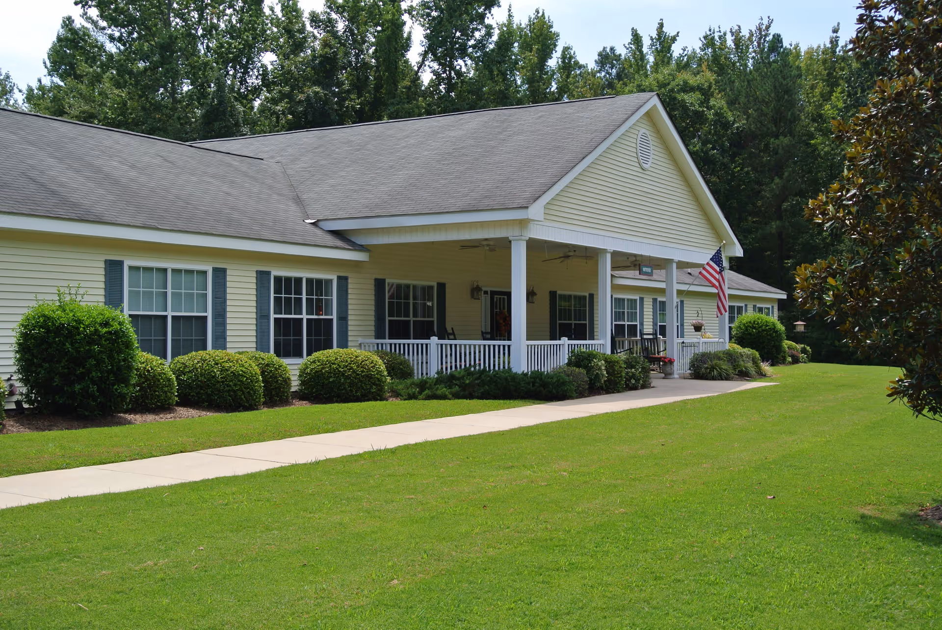Single-story yellow building with a covered porch, white railing, and several windows. The porch has rocking chairs and an American flag hanging near the entrance. The building is surrounded by neatly trimmed bushes and a well-maintained green lawn with a concrete walkway leading to the porch. Trees are visible in the background.