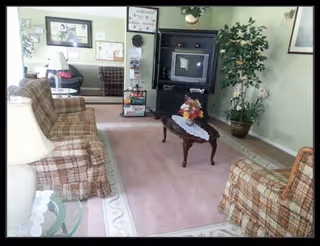 Cozy living room with plaid armchairs, a coffee table topped with a floral arrangement, an entertainment cabinet with a TV, and potted plants.