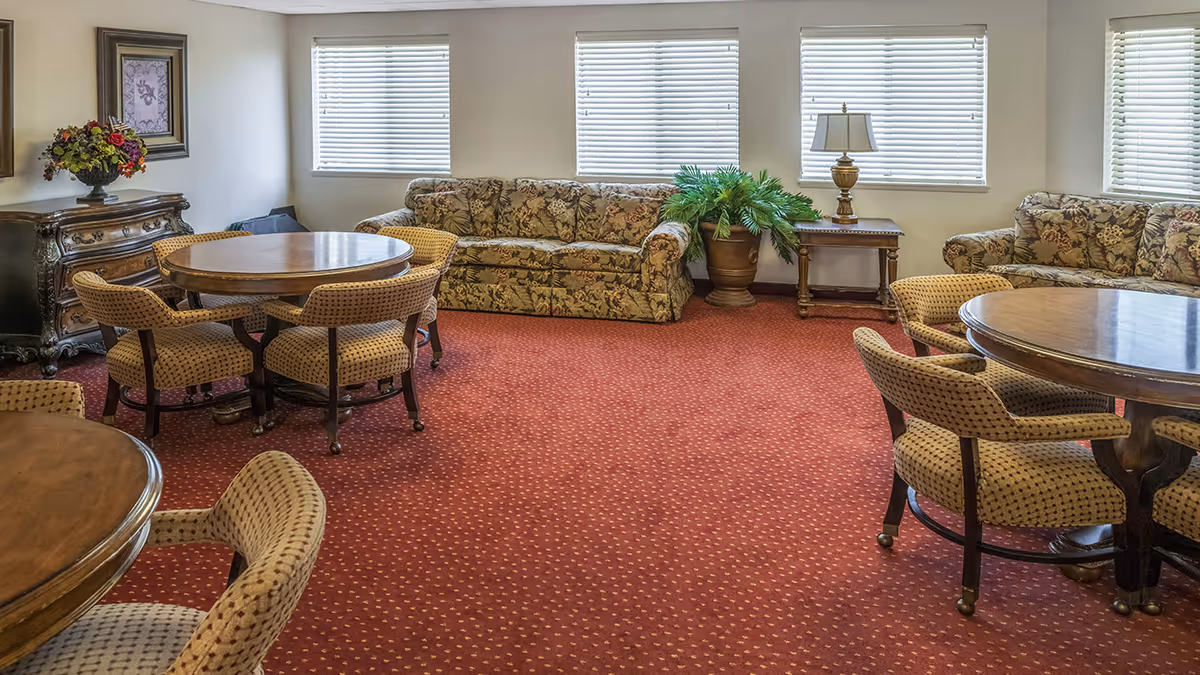 A senior living common room with round wooden tables and upholstered chairs, floral-patterned sofas, a potted plant, and windows with blinds.