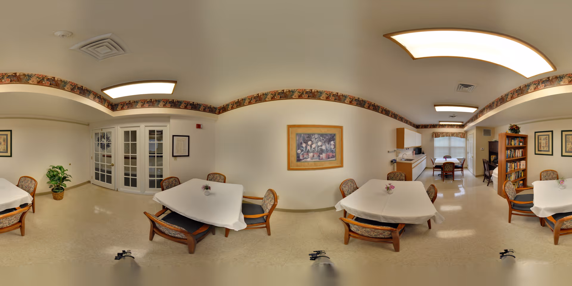 A well-lit common area in a senior living facility with multiple tables covered with white tablecloths and surrounded by wooden chairs with patterned cushions. The room features a decorative border near the ceiling, framed artwork on the walls, a potted plant, a bookshelf filled with books, and a kitchenette area with cabinets and a sink. The floor is light-colored and the ceiling has recessed lighting panels.