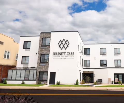 Exterior view of a modern three-story building with white and stone facade, large windows, and a sign that reads 'Serenity Care Personal Care Community Old Forge'. The building is set against a partly cloudy sky and has a small lawn and sidewalk in front.