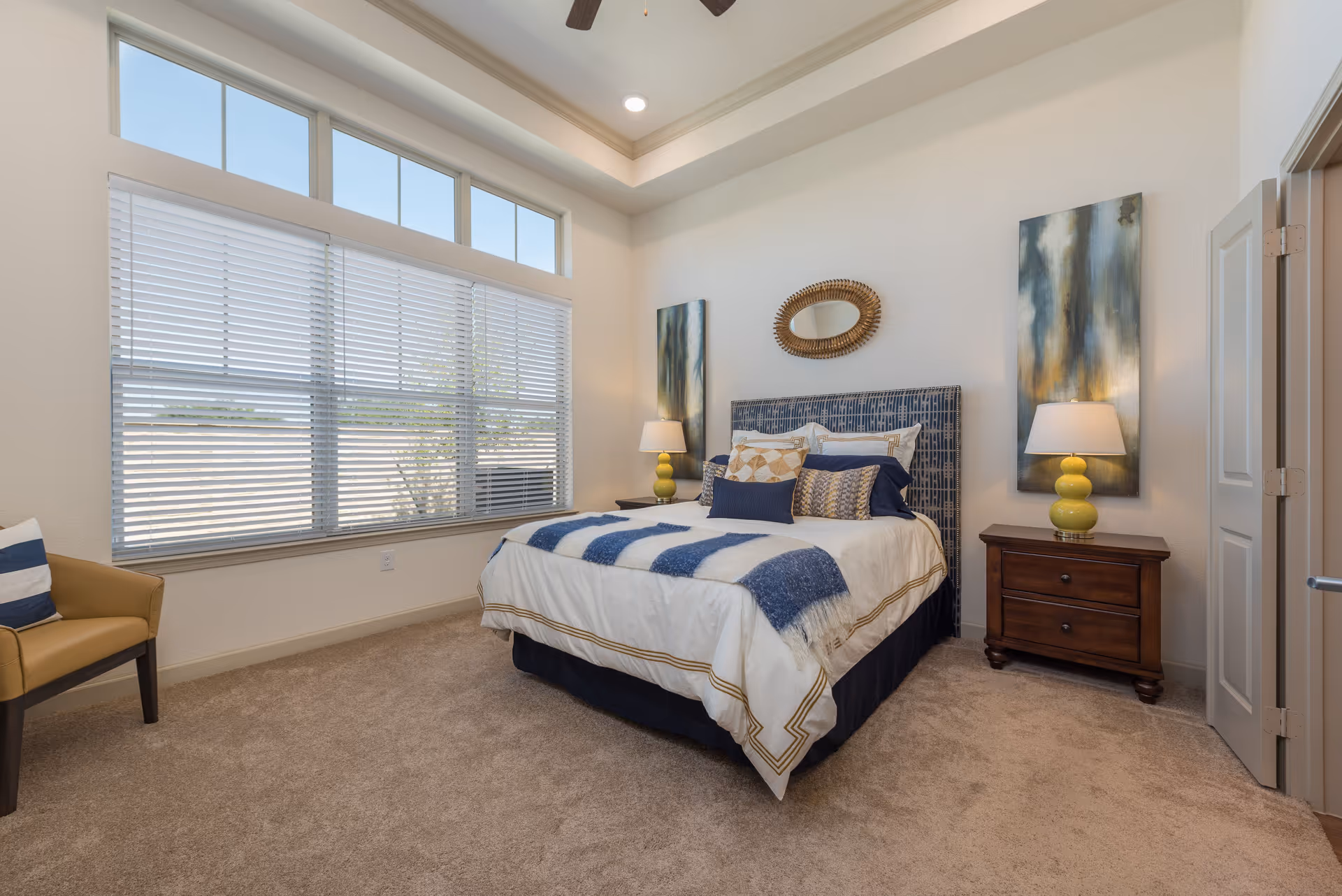 A bright bedroom with a large window covered by white blinds. The room features a bed with white and blue bedding, two bedside tables with yellow lamps, abstract artwork on the walls, a decorative mirror above the bed, and a beige armchair with a striped pillow in the corner.