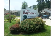 A white sign with dark red and black text reading 'The Bayberry Retirement Inn' surrounded by green bushes and grass, with residential buildings and a parked car visible in the background under a partly cloudy sky.