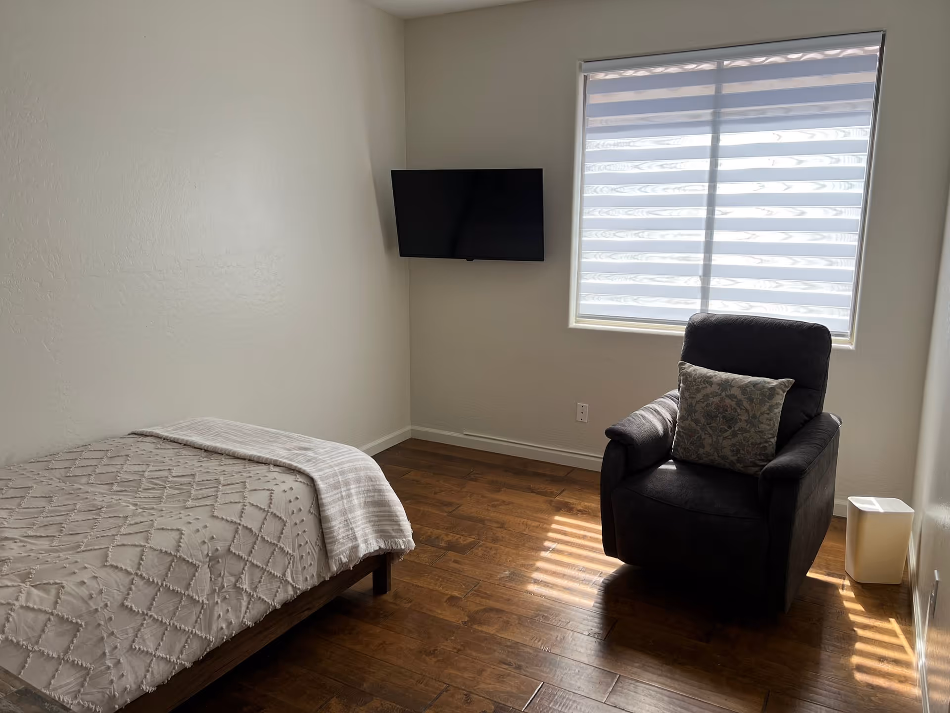 Small bedroom with a single bed, a reclining armchair, a wall-mounted TV, and a window with blinds.