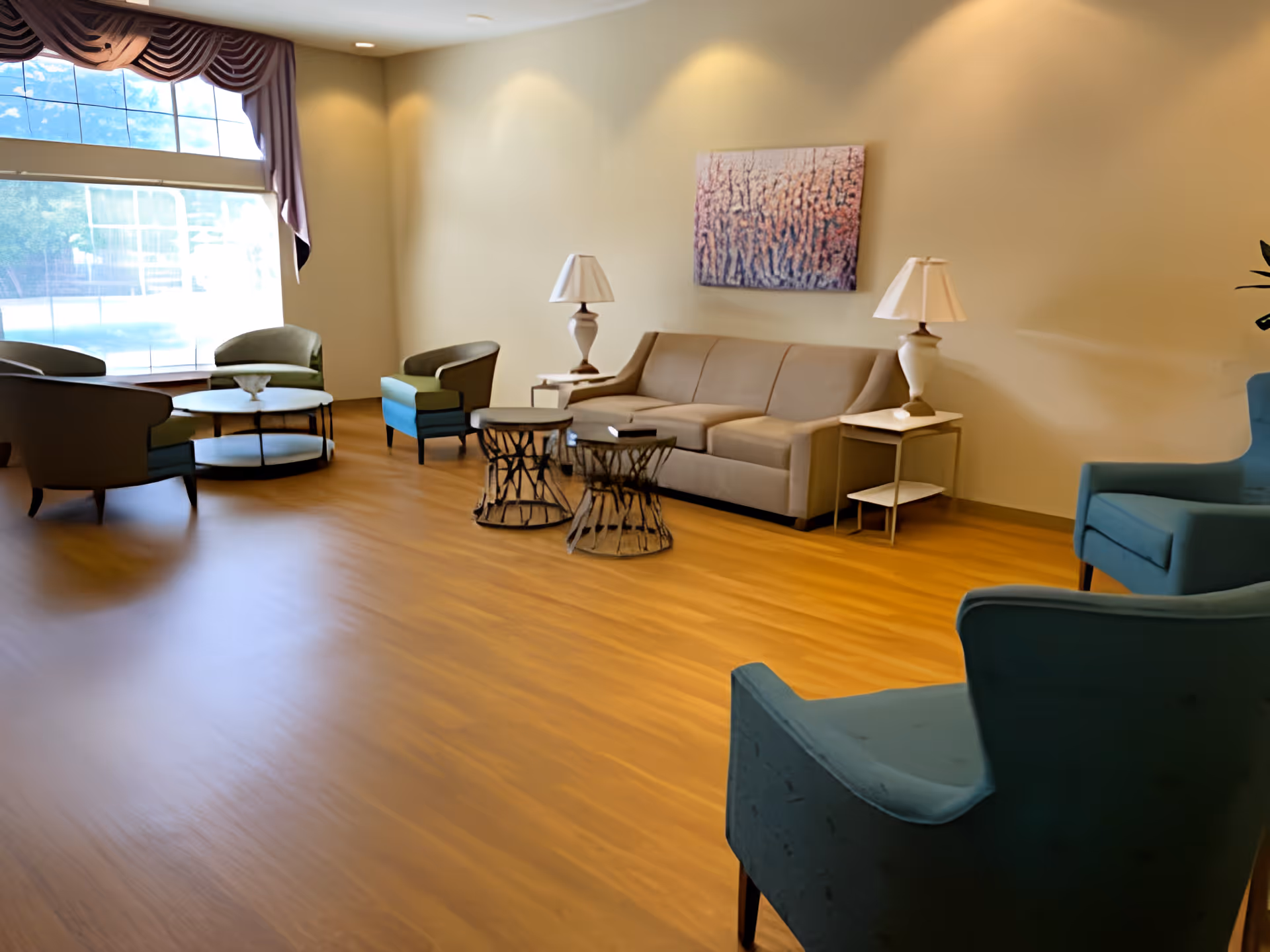 A spacious living room with wooden flooring, featuring a beige sofa with two side tables and lamps, two blue armchairs in the foreground, and a seating area with green chairs and a round coffee table near a large window with purple curtains. A colorful abstract painting hangs on the wall above the sofa.