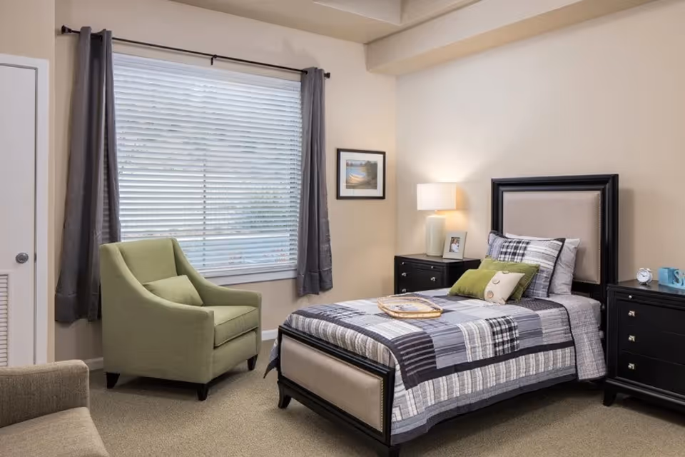 A neatly arranged bedroom with a single bed featuring a black and beige headboard and a patchwork quilt in shades of gray, white, and black. There are two green accent pillows and one beige pillow on the bed. A wooden tray is placed on the bed. To the right of the bed is a black nightstand with a white lamp, a framed photo, and a small clock. Another black dresser is visible with a blue decorative item on top. A green armchair with a matching pillow is positioned near a window with closed blinds and gray curtains. The walls are painted beige, and a framed picture hangs on the wall beside the bed.