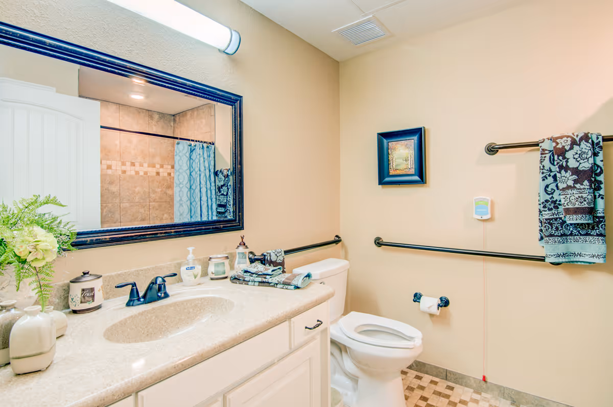 A clean and well-lit bathroom featuring a white vanity with a beige countertop and an oval sink. On the countertop are decorative items including a small plant, soap dispenser, and candles. Above the vanity is a large mirror with a dark frame and a horizontal light fixture. The bathroom has beige walls, a white toilet with safety grab bars on the walls, a towel rack with a patterned towel, and a small framed picture on the wall. The shower area with tiled walls and a patterned shower curtain is visible in the mirror reflection.