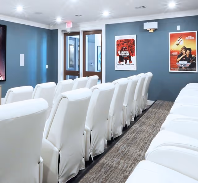 Interior view of a small theater room with rows of white cushioned chairs facing a screen. The walls are painted gray and decorated with framed movie posters, including one for Top Gun. There is a carpeted aisle between the rows of chairs and a glass door at the back of the room.