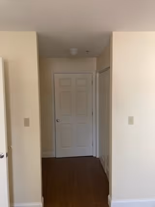 A hallway with beige walls and wooden flooring leading to a white door at the end. There is a closet door on the right side of the hallway and light switches on the walls.
