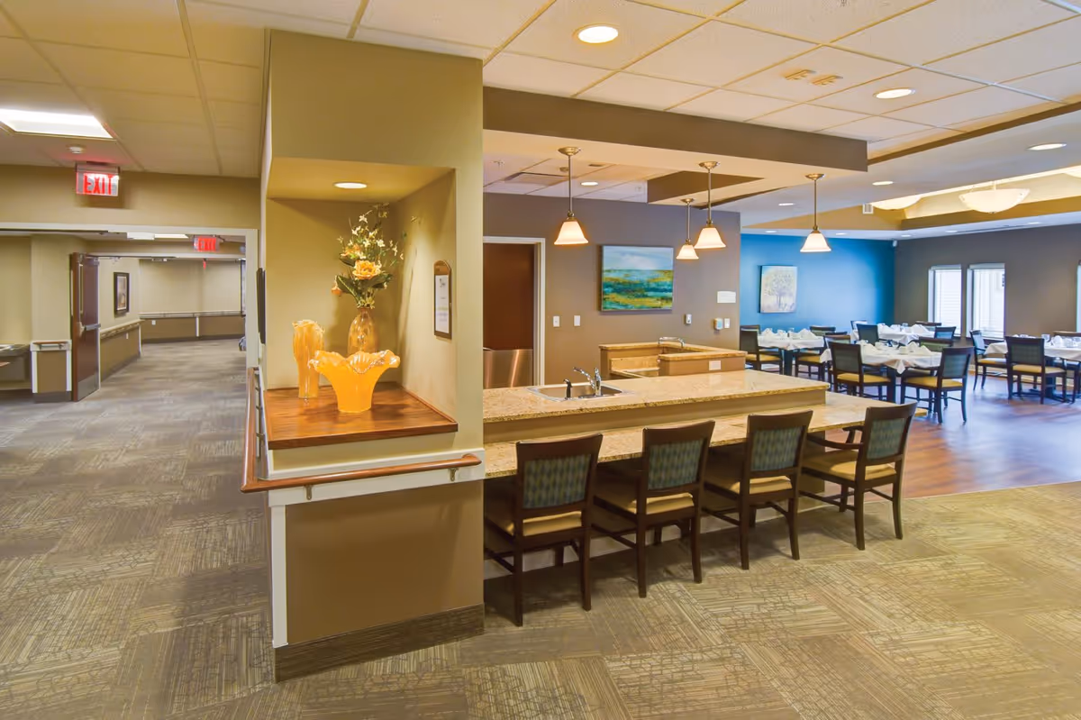 Open communal dining area with a long granite counter and barstools, dining tables set for meals, and a decorative vase on a ledge.