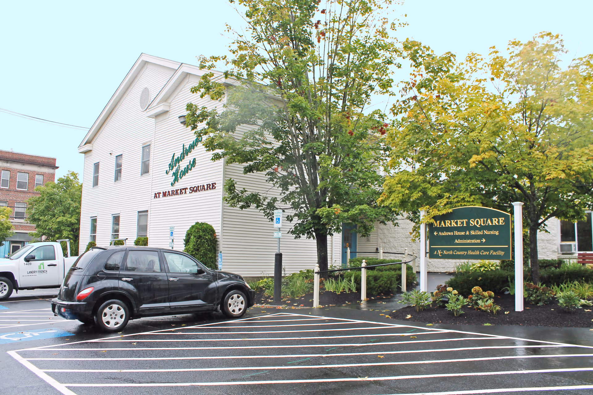 Exterior view of a senior living facility named Andrews House at Market Square, showing a two-story white building with green and red signage. There are trees and shrubs around the building, a parking lot with a black car and a white truck, and a green and yellow sign indicating Market Square with directions to Andrews House & Skilled Nursing, Administration, and North Country Health Care Facility.