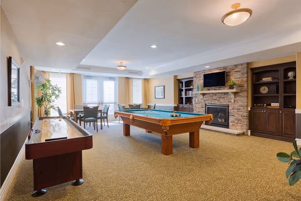 A well-lit recreational room featuring a pool table with balls arranged on it, a shuffleboard table, several tables with chairs near large windows with curtains, a stone fireplace with a mounted TV above it, and built-in wooden shelves with decorative items.