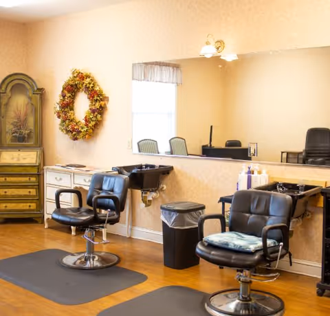 Interior of a salon area in a senior living facility with two black salon chairs in front of sinks for hair washing. There is a large mirror on the wall, a decorative wreath, a wooden cabinet, and a trash bin. The room has light-colored wallpaper and wooden flooring.
