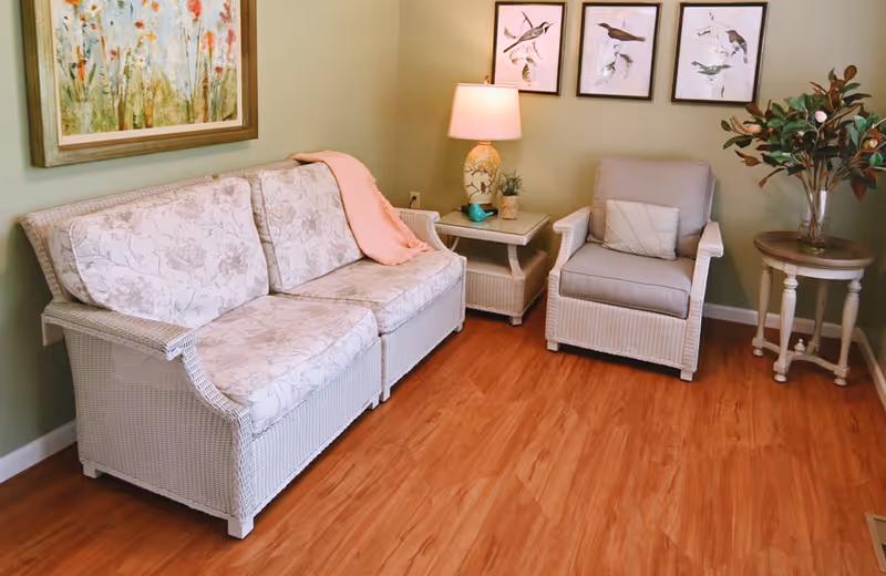 A cozy sitting area with a white wicker loveseat and armchair, both with floral cushions. A pink throw blanket is draped over the loveseat. Between the chairs is a small wicker side table with a decorative lamp and small plant. On the right side, there is a round wooden table with a vase of greenery. The walls are light green and decorated with framed artwork featuring birds and floral designs. The floor is a warm wood tone.