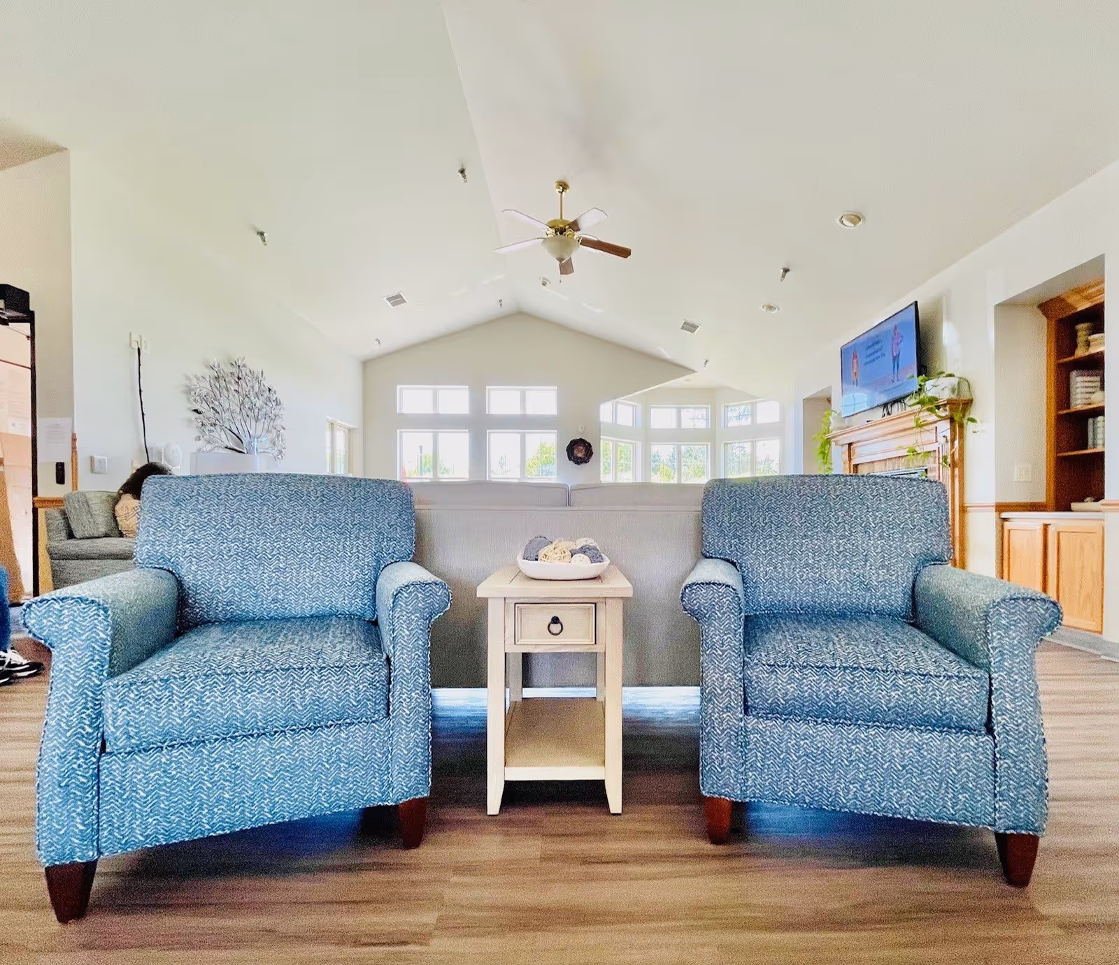 A bright and spacious living room area with two blue patterned armchairs facing the camera, separated by a small wooden side table with decorative items. Behind the chairs is a light gray sofa, large windows letting in natural light, a ceiling fan, a wall-mounted TV above a wooden fireplace mantel, and built-in wooden shelves on the right.